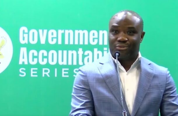 Speaker at a podium wearing a light blue blazer speaks into a microphone, with a green backdrop that reads 'Government Accountability Series'.
