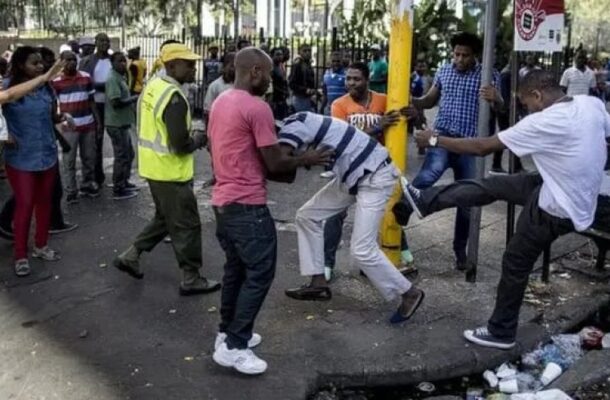 image_870x_6a30fe0fa23c5 | Ghanaguardian.com Crowd of onlookers watches as several men restrain a man in a striped shirt near a yellow pole on a city street, debris on the ground nearby.