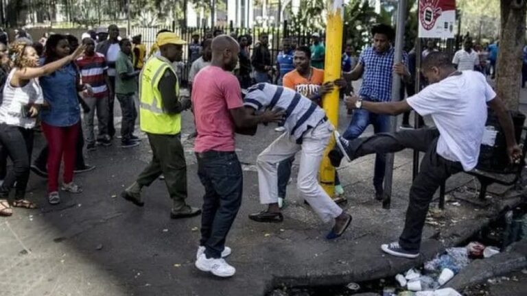 Crowd of onlookers watches as several men restrain a man in a striped shirt near a yellow pole on a city street, debris on the ground nearby.