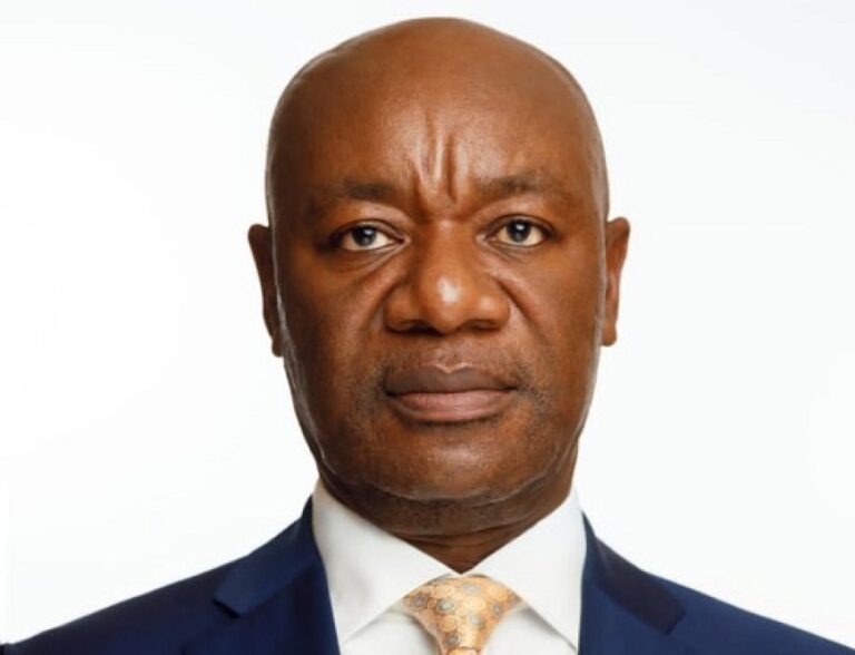 Portrait of a bald Black man in a navy suit and peach tie, looking at the camera with a neutral expression.