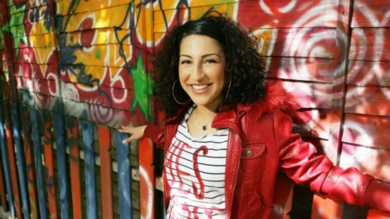 Smiling woman with curly dark hair in a red jacket leaning on a colorful graffiti-covered fence/wall backdrop.