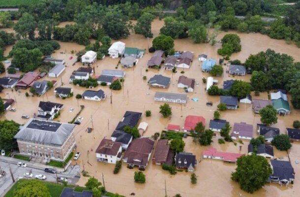 Aerial view of a residential town nearly submerged in brown floodwater, with streets, yards, and cars visible beneath the waterline.
