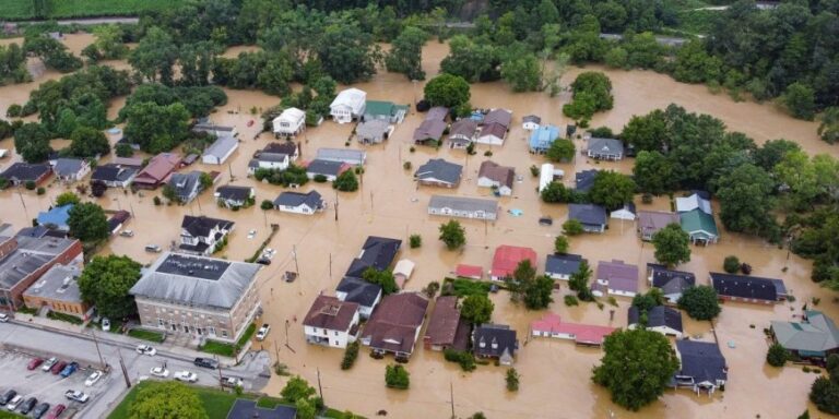 Aerial view of a residential town nearly submerged in brown floodwater, with streets, yards, and cars visible beneath the waterline.