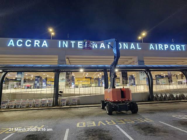 Night view of Accra International Airport with a mobile elevating work platform in the foreground near the terminal entrance.