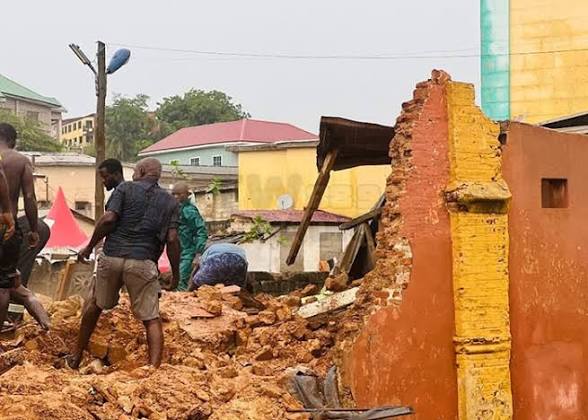Group of men standing amid rubble and a partially collapsed brick wall in an urban street, with damaged buildings in the background.