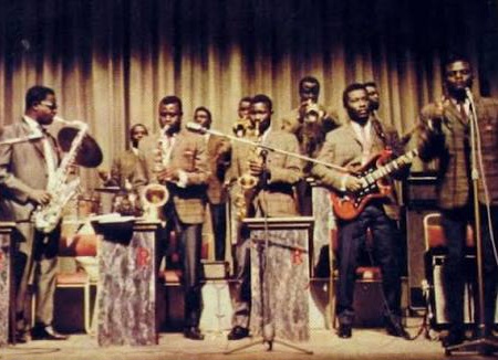 A group of male musicians in suits performing on a stage, playing saxophones, trumpets, and a guitar in a vintage jazz ensemble