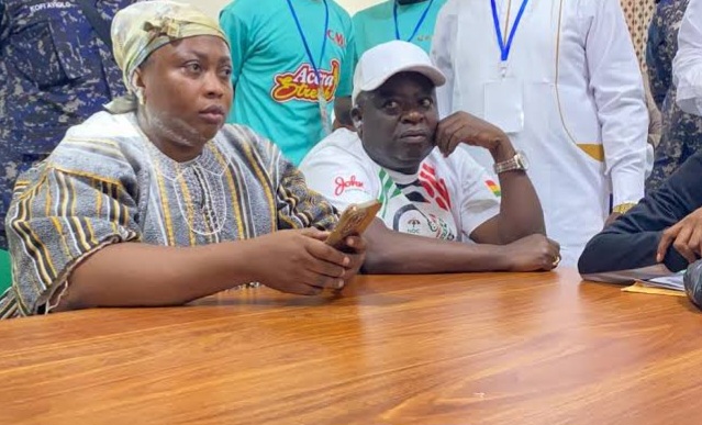 Two people seated at a wooden table during a meeting; the woman on the left holds a smartphone while the man beside her rests his chin on his hand, with others standing in the background.