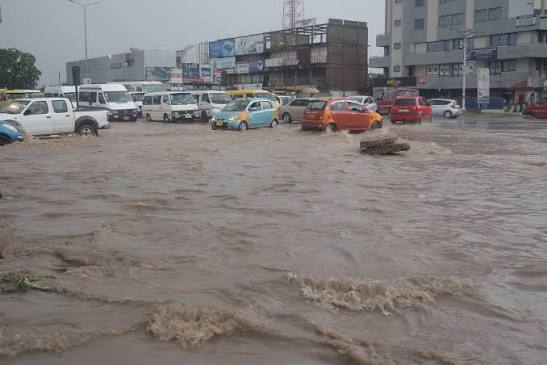 Urban street submerged by floodwaters; cars and vans stranded as brown water covers the road near storefronts and buildings.