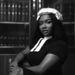 Black-and-white portrait of a Black woman wearing a white era wig and dark dress with a white collar, seated with arms crossed in front of a bookshelf filled with legal volumes.