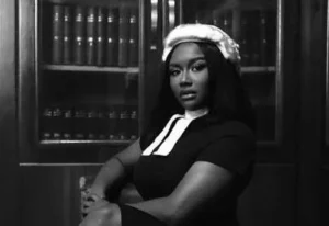 Black-and-white portrait of a Black woman wearing a white era wig and dark dress with a white collar, seated with arms crossed in front of a bookshelf filled with legal volumes.