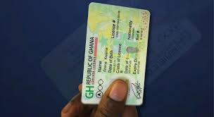 images | Ghanaguardian.com Person holding a green Ghana national ID card with photo and hologram visible on a blue background.