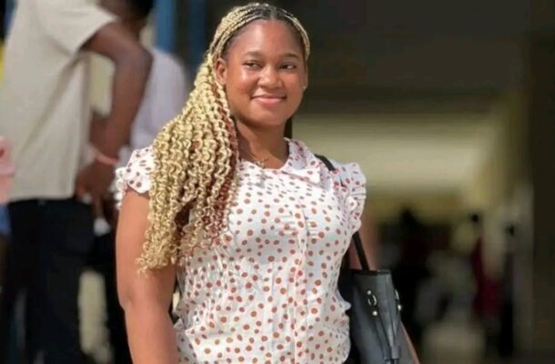 Smiling woman with blonde box braids walking outdoors, wearing a white polka-dot blouse and carrying a black bag.