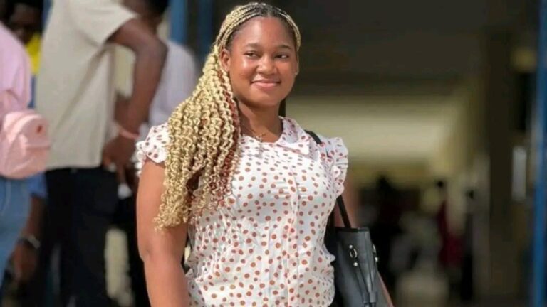 Smiling woman with blonde box braids walking outdoors, wearing a white polka-dot blouse and carrying a black bag.