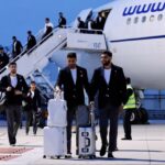 Group of men in suits pulling luggage away from a white passenger plane on the tarmac, stairs and ground crew nearby.
