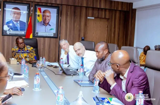 jinapor | Ghanaguardian.com Group of six men in a formal meeting around a long conference table with laptops and water bottles in a wood-paneled room; two framed portraits and a flag on the wall, Ministry of Energy and Transformation branding nearby.