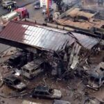 Aerial view of a collapsed building with burned cars and debris, firefighters and responders on a wet street at a disaster site.