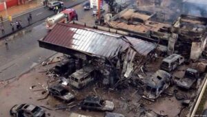 Aerial view of a collapsed building with burned cars and debris, firefighters and responders on a wet street at a disaster site.