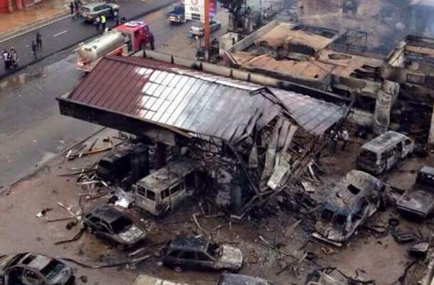 Aerial view of a collapsed building with burned cars and debris, firefighters and responders on a wet street at a disaster site.