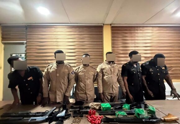 Six uniformed individuals with blurred faces stand behind a table displaying multiple rifles, magazines, and red ammunition cartridges in a room with wooden blinds.