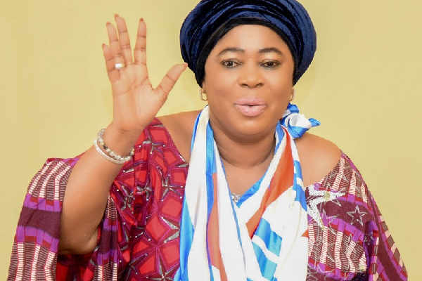 Woman wearing a colorful traditional outfit and blue headwrap raises her hand as if greeting.