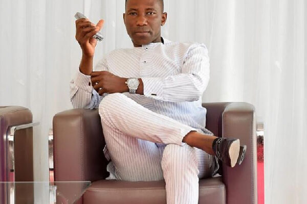 Man in a light striped suit sits cross‑legged in a brown chair, holding a remote control.