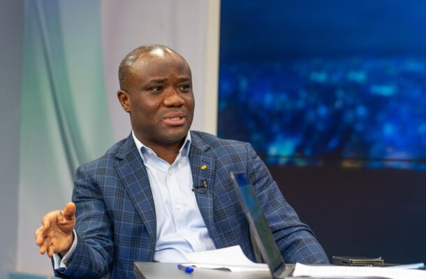 Man in a blue check blazer speaks while seated at a news desk with a laptop and papers in a studio setting.