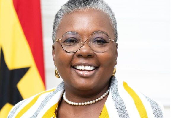 madam | Ghanaguardian.com Smiling Black woman with short gray hair, round glasses, pearl necklace, and a yellow-and-white striped scarf; flag in the background.