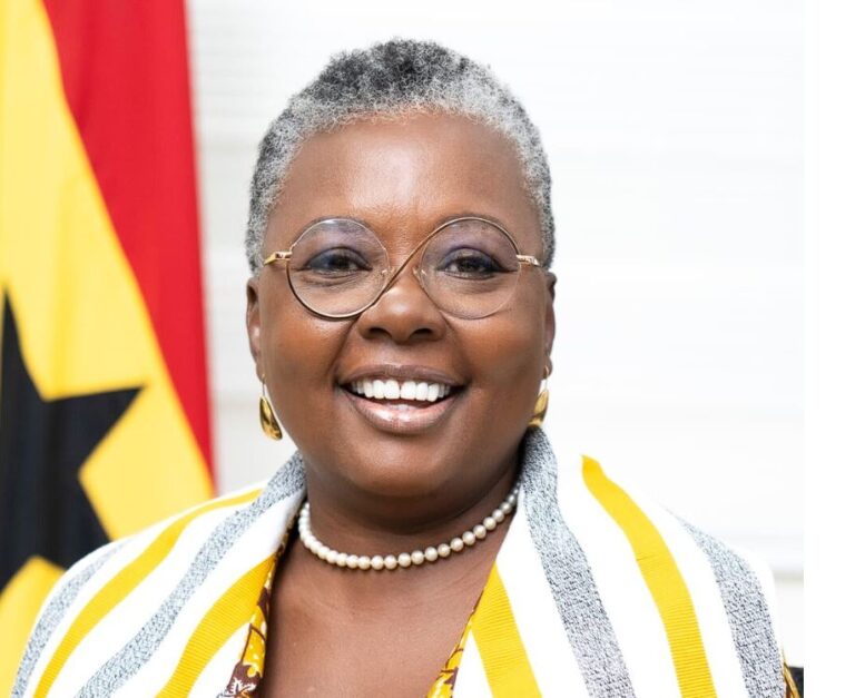 Smiling Black woman with short gray hair, round glasses, pearl necklace, and a yellow-and-white striped scarf; flag in the background.