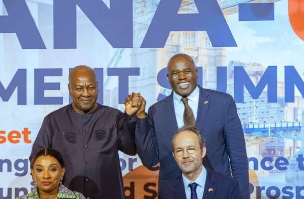 Four people pose on a conference stage; two men stand and clasp hands, a seated man and woman smile in front of a large blue backdrop with bold lettering