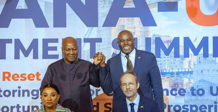 Four people pose on a conference stage; two men stand and clasp hands, a seated man and woman smile in front of a large blue backdrop with bold lettering
