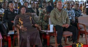 A formal ceremony with a woman in a brown dress and a man in an olive shirt seated in front, with uniformed officers and a crowd behind them.