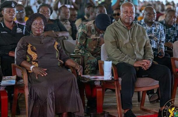 A formal ceremony with a woman in a brown dress and a man in an olive shirt seated in front, with uniformed officers and a crowd behind them.