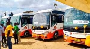 Row of white-and-orange Metro Mass Transit buses parked on a dusty lot, with people standing nearby at what appears to a launch event.