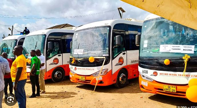Row of white-and-orange Metro Mass Transit buses parked on a dusty lot, with people standing nearby at what appears to a launch event.