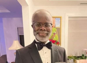 Smiling older man in a black tuxedo, white shirt, and bow tie, wearing round glasses, indoors at a social event.