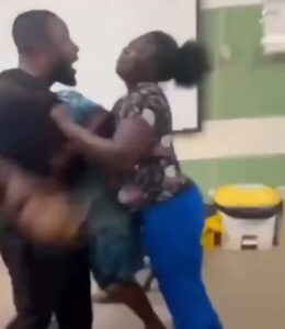 Two adults appear to be in a physical struggle indoors, grappling with each other; a bearded man faces a woman in a patterned top and blue pants, with a yellow-and-white cooler in the background.
