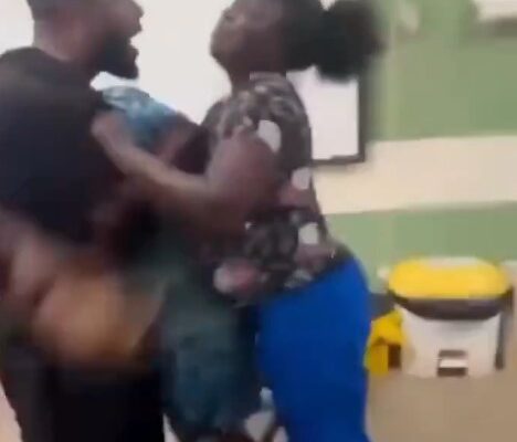 Two adults appear to be in a physical struggle indoors, grappling with each other; a bearded man faces a woman in a patterned top and blue pants, with a yellow-and-white cooler in the background.