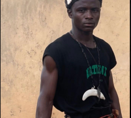 Young man standing outdoors in a black T-shirt, wearing a patterned head wrap and a white horn-shaped pendant with a rope belt.