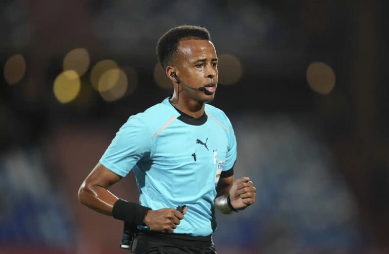 Soccer referee in a light blue uniform with a headset mic, running on the field during a match