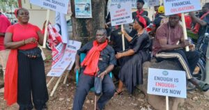 Group of protesters, some seated and others standing, holding signs with messages about unpaid debts and workers’ rights; one sign reads 'Enough is enough - Pay us now'.