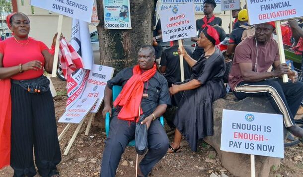 Group of protesters, some seated and others standing, holding signs with messages about unpaid debts and workers’ rights; one sign reads 'Enough is enough - Pay us now'.