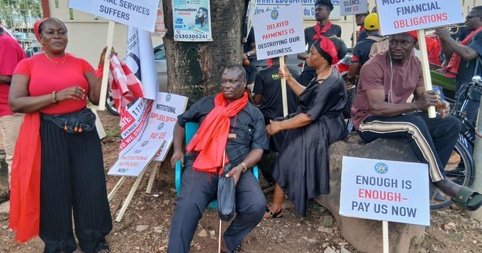 Group of protesters, some seated and others standing, holding signs with messages about unpaid debts and workers’ rights; one sign reads 'Enough is enough - Pay us now'.