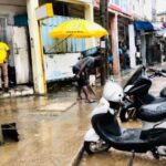 Street scene with flooded sidewalk, two scooters parked in foreground, and people wading near shops under colorful awnings.