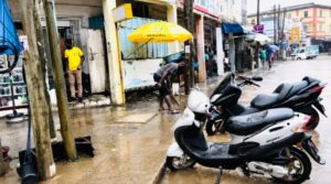 Street scene with flooded sidewalk, two scooters parked in foreground, and people wading near shops under colorful awnings.