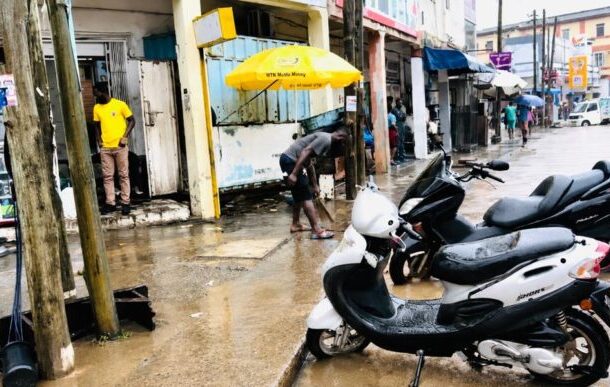takoradi-696x387 | Ghanaguardian.com Street scene with flooded sidewalk, two scooters parked in foreground, and people wading near shops under colorful awnings.