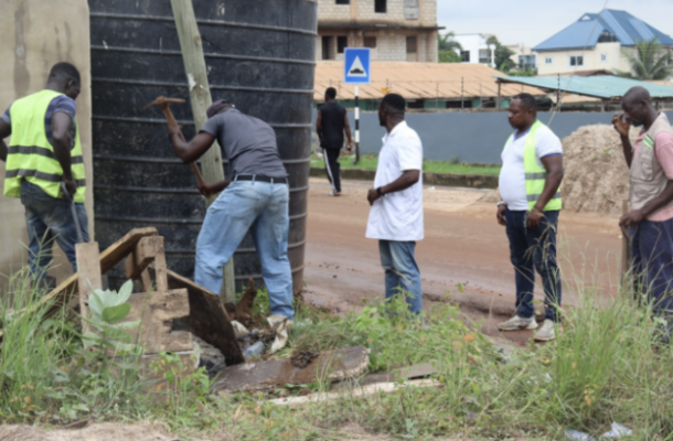 Workers in safety vests repair a water storage tank at a roadside site, with others observing nearby on a dirt road.