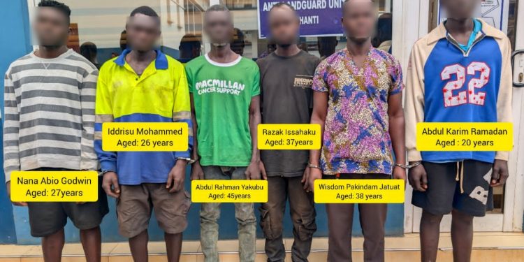 Seven men stand in a row in front of a shop, each with a bright yellow tag showing a name and age in bold text (blurred faces for privacy).