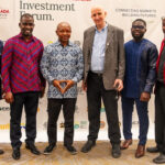Six men in smart casual attire posing for a photo at a Ghana Investment Forum event, standing in front of a banner with logos.