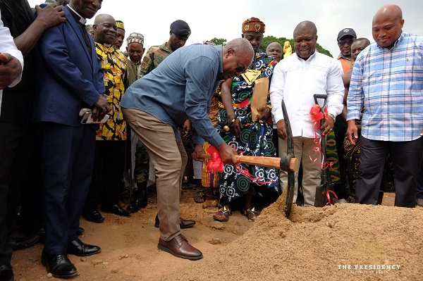 Man in a blue shirt uses a ceremonial hammer to break ground at a formal groundbreaking ceremony, surrounded by officials and onlookers on a dirt site.