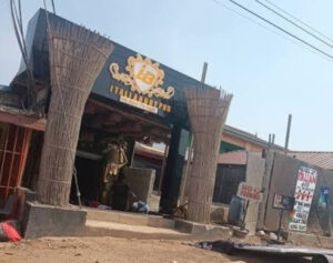 Open-front rustic cafe/restaurant with straw-wrapped pillars and a crest sign above the entrance; sandy ground and power lines overhead.